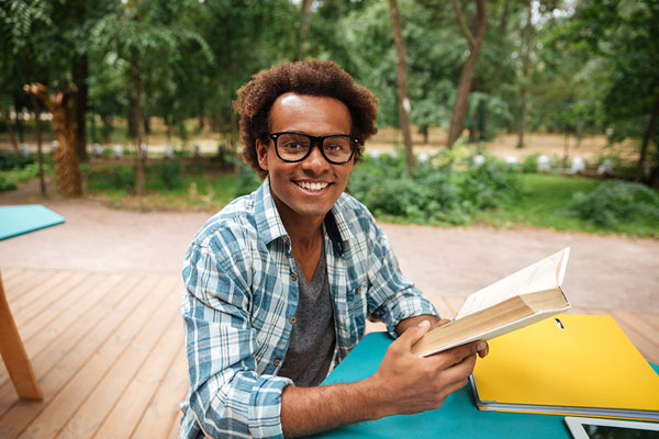Young man with glasses smiling and reading outdoors